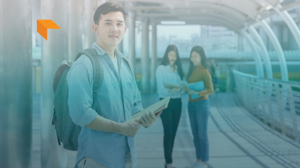 A young East Asian male student wearing a light blue denim shirt and backpack, holding books and looking directly at the camera with a subtle smile. In the blurred background, two female students stand together on a modern covered pedestrian bridge or walkway. An orange stylized arrow logo appears in the top-left corner, and a blue-to-light gradient overlay spans across the image.