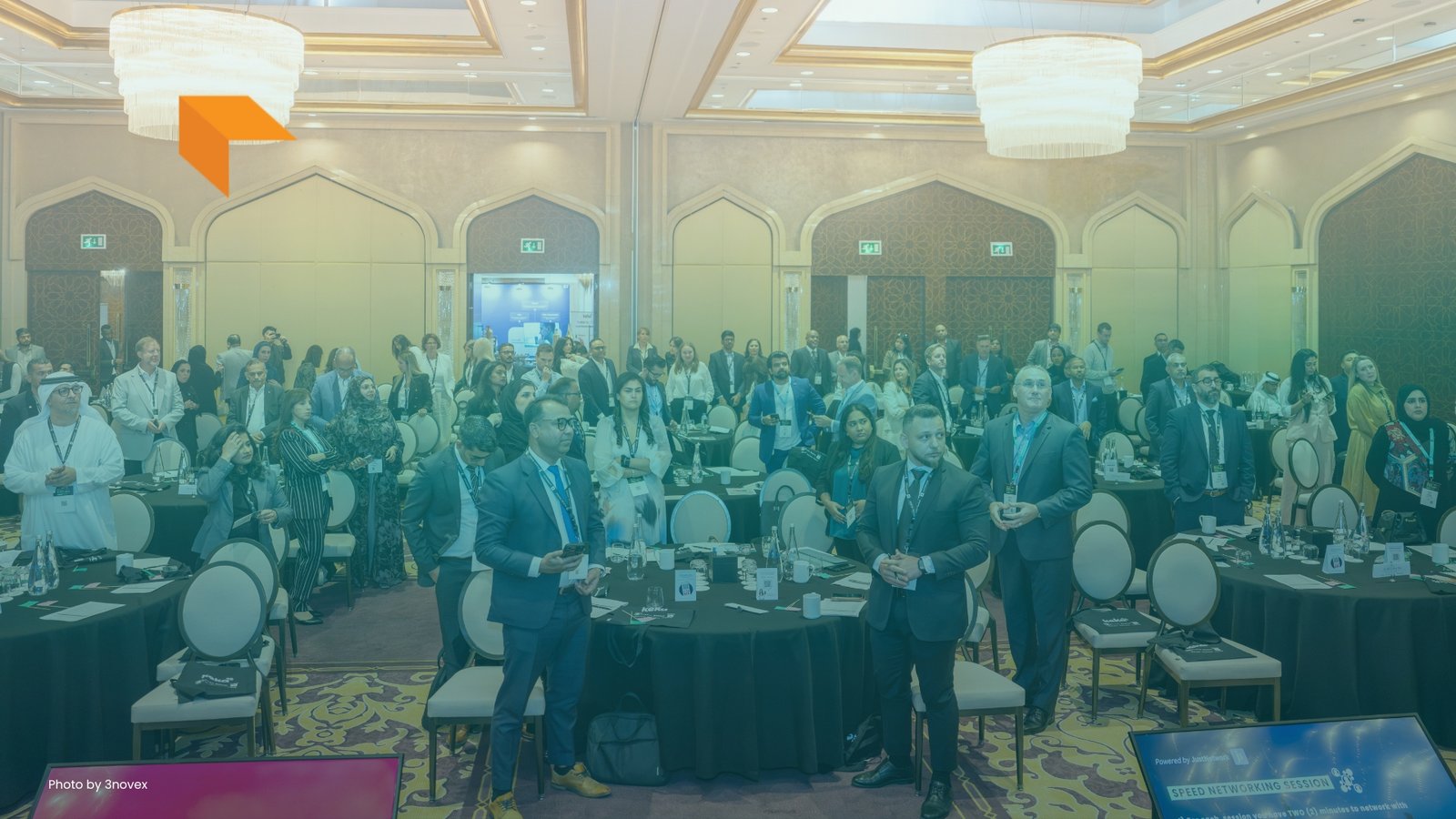 A large group of attendees standing around round tables in an elegant banquet hall during a networking event, with tiered chandeliers above and an orange logo graphic in the upper-left corner.